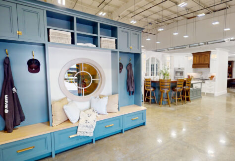 Virtual kitchen showroom featuring a blue built-in mudroom storage unit in the foreground and multiple kitchen displays with cabinetry, an island, and bar seating in a bright, open showroom space.